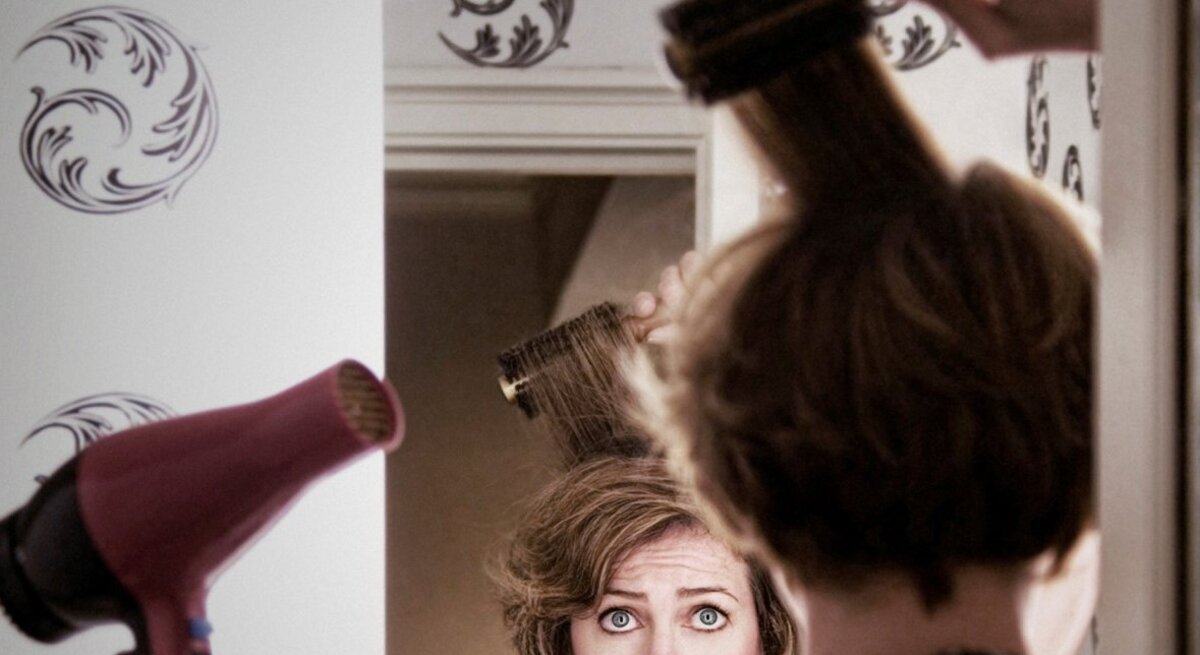 Eine Frau mit kurzen braunen Haaren schaut ernst in den Spiegel, während sie mit Rundbürste und Föhn ihre Haare stylt.