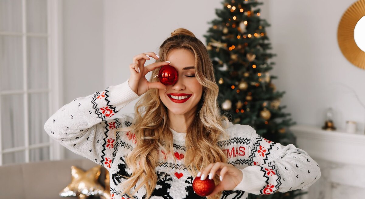 Junge Frau mit roten Lippen und Weihnachtspullover posiert fröhlich mit Weihnachtskugeln vor einem dekorierten Baum.