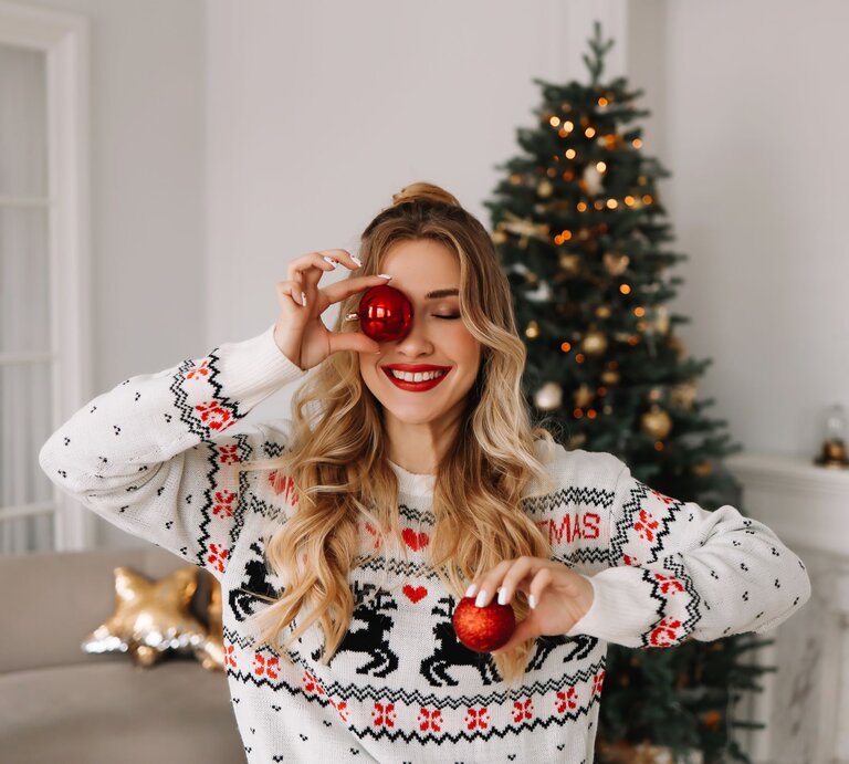Junge Frau mit roten Lippen und Weihnachtspullover posiert fröhlich mit Weihnachtskugeln vor einem dekorierten Baum. | © Getty Images/ Elizaveta Starkova