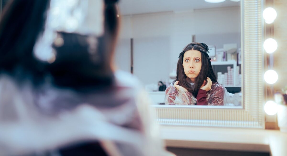 Frau im Frisiersalon, die besorgt oder erschrocken in den Spiegel schaut. Sie hat Frisiurzubehör im Haar und trägt einen Frisierumhang. Ihr Gesichtsausdruck deutet darauf hin, dass sie über das Ergebnis des Haarschnitts nicht glücklich ist. 