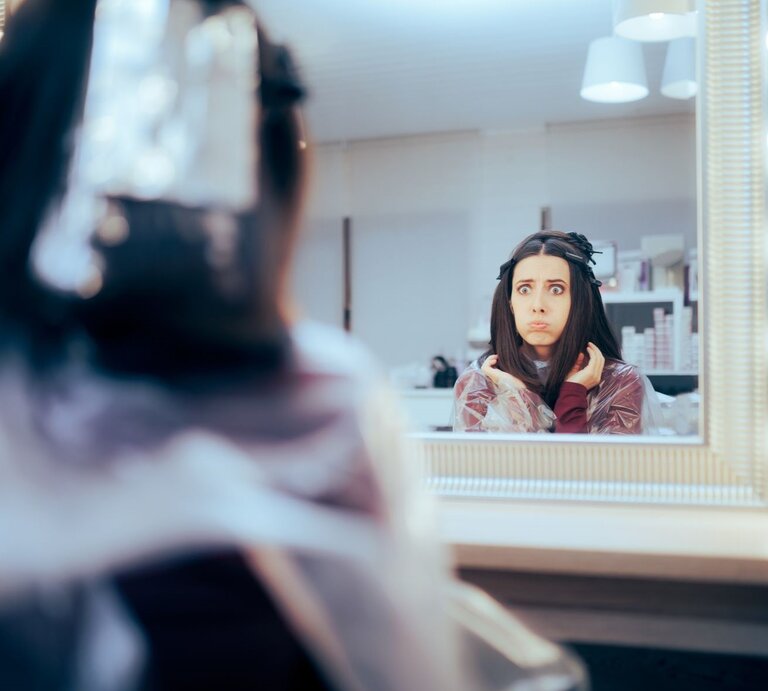 Frau im Frisiersalon, die besorgt oder erschrocken in den Spiegel schaut. Sie hat Frisiurzubehör im Haar und trägt einen Frisierumhang. Ihr Gesichtsausdruck deutet darauf hin, dass sie über das Ergebnis des Haarschnitts nicht glücklich ist.  | © Getty Images / nicoletaionescu