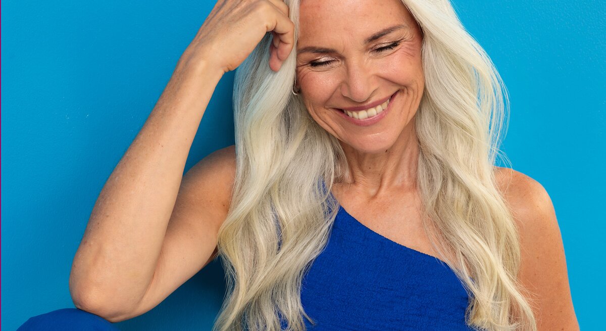 Lachende Frau im mittleren Alter mit langen grauen Haaren und blauem Oberteil lehnt sich entspannt an eine blaue Wand und blickt lächelnd nach unten.