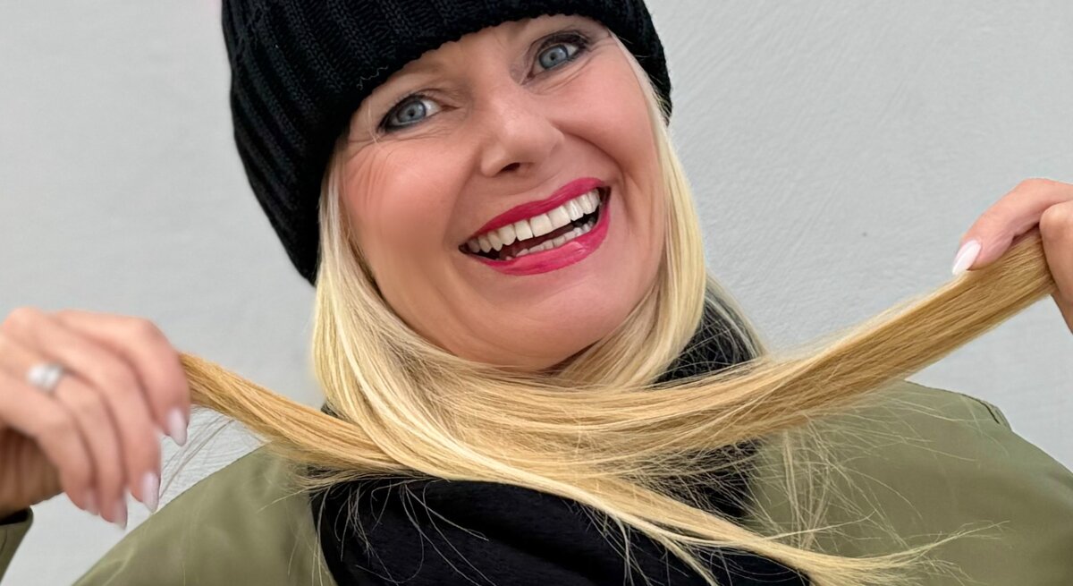 Fröhlich lächelnde Frau mit blonden Haaren, die eine schwarze Strickmütze trägt. Sie hält ihre Haare in den Händen und präsentiert sie lachend in die Kamera.