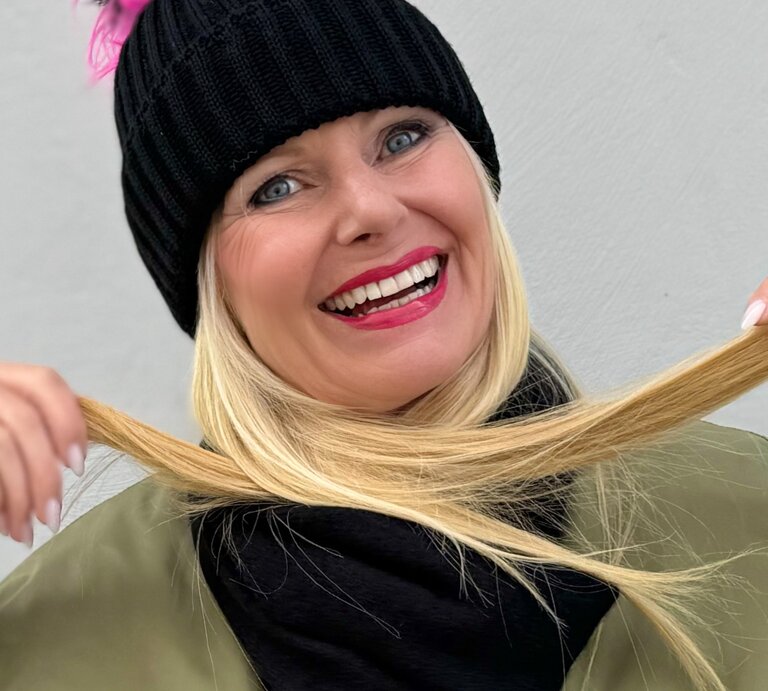 Fröhlich lächelnde Frau mit blonden Haaren, die eine schwarze Strickmütze trägt. Sie hält ihre Haare in den Händen und präsentiert sie lachend in die Kamera. | © Renate Zott