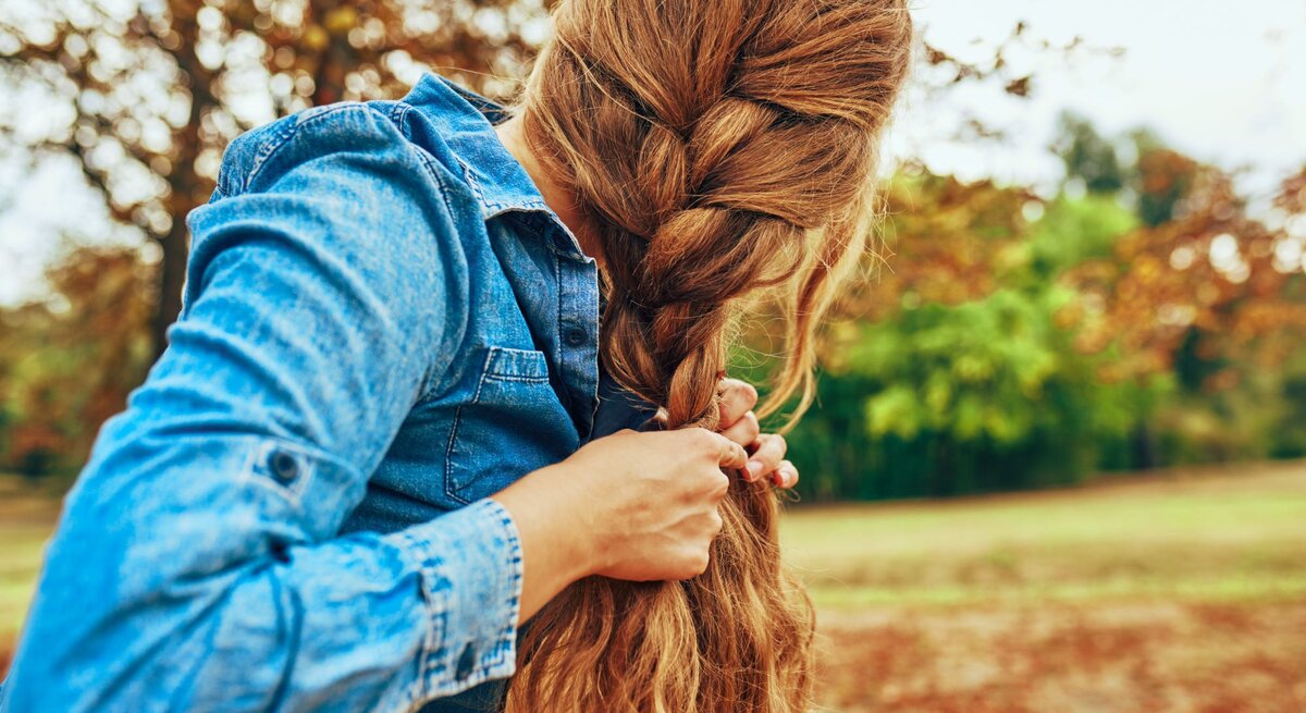 Zu sehen ist eine Frau in der Natur, die sich die Haare zu einem französischen Zopf flechtet