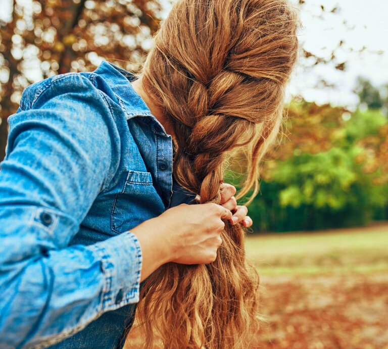 Zu sehen ist eine Frau in der Natur, die sich die Haare zu einem französischen Zopf flechtet | © Shutterstock/Yuricazac