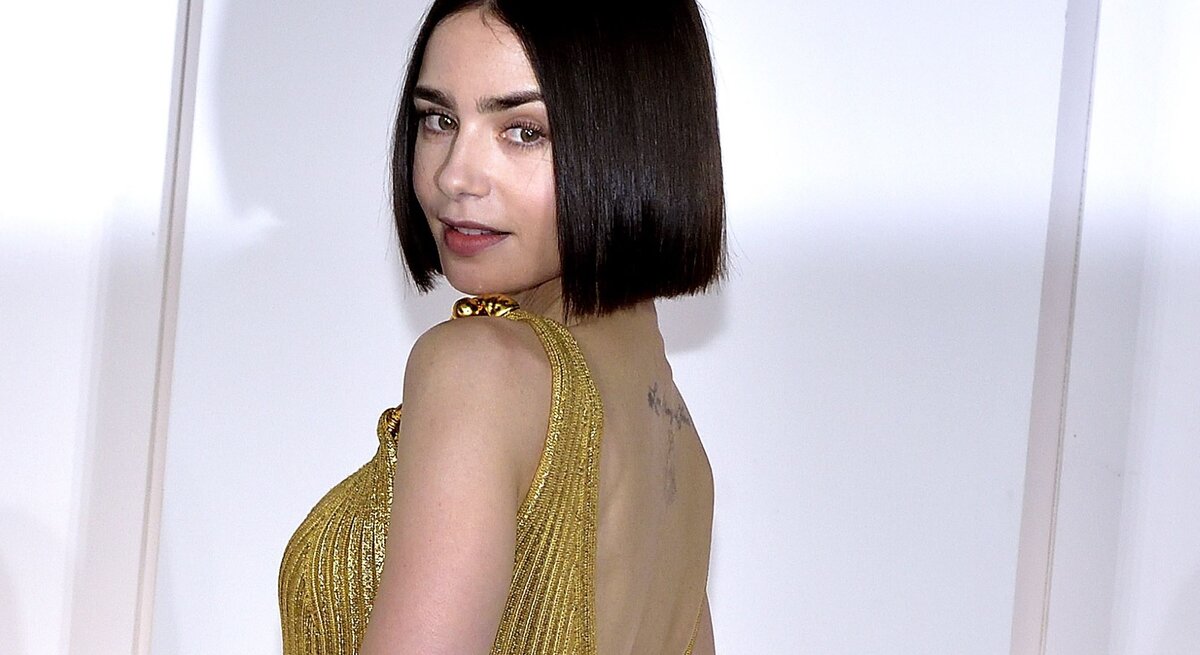 Frau mit einem bob-geschnittenen, dunklen Haar, die eine elegante, goldene Robe trägt. Sie steht mit dem Rücken zur Kamera und dreht sich über die Schulter, wobei sie direkt in die Kamera blickt.
