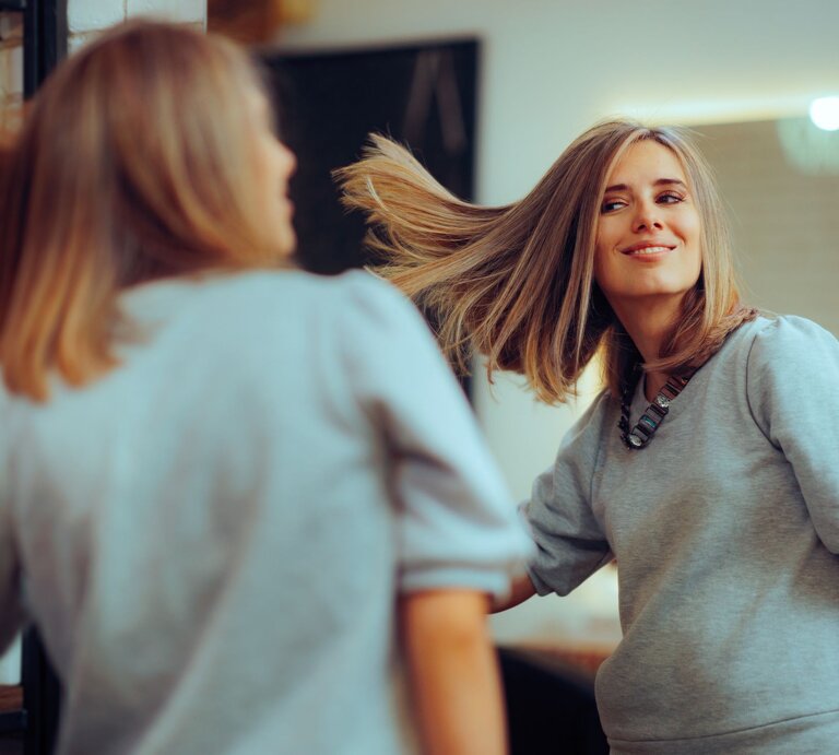 Junge Frau, die sich in einem Spiegel betrachtet und dabei spielerisch ihr Haar schwingt. Sie trägt ein lässiges graues T-Shirt und eine modische Halskette. Ihre Haare sind zu einem schicken Long Bob geschnitten. | © Adobe Stock / nicoletaionescu