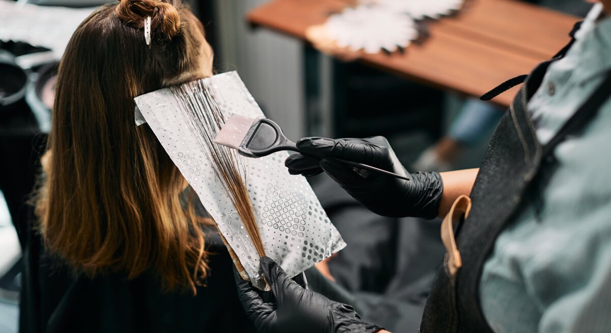 Friseur mit schwarzem Handschuh trägt mit Pinsel Haarfarbe auf braune Strähnen einer Frau auf, die in einem Salon sitzt.