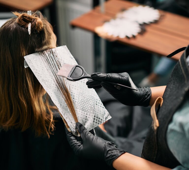 Friseur mit schwarzem Handschuh trägt mit Pinsel Haarfarbe auf braune Strähnen einer Frau auf, die in einem Salon sitzt. | © Adobe Stock/ Drazen