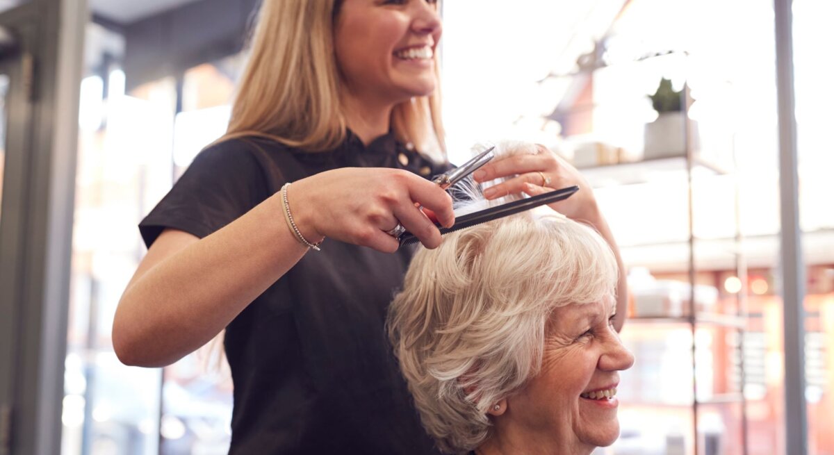 Eine junge Friseurin mit langen blonden Haaren schneidet lächelnd die Haare einer fröhlichen älteren Dame in einem hellen Salon.