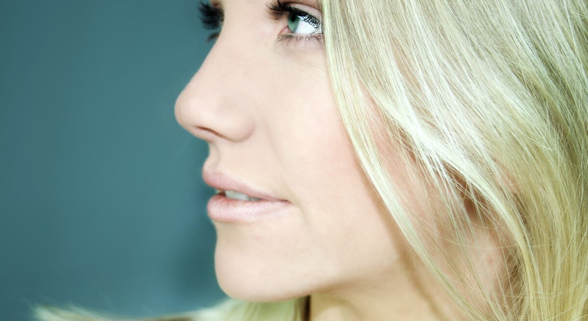 Frau im Profil mit langen, blonden Haaren, die einen Grünstich haben. Ihre Haare sind glatt und fallen sanft über ihre Schulter. Sie hat einen intensiven Blick