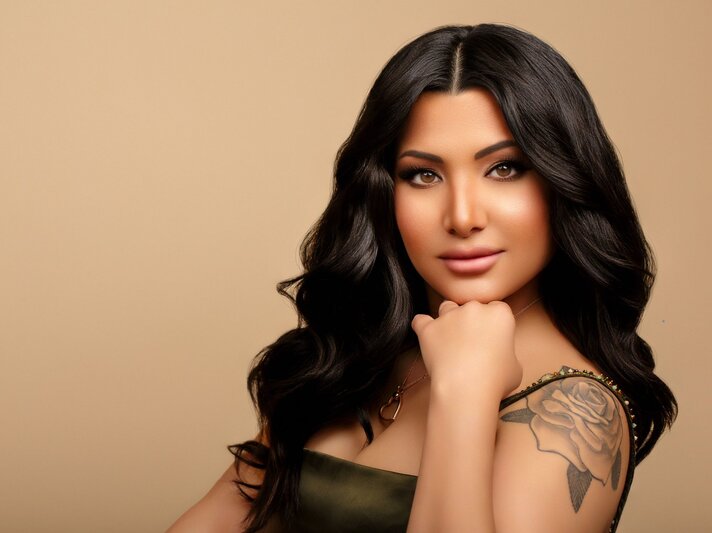 Frisörin Nesrin Obermeier mit langen, gewellten, dunklen Haaren und Make-up. Sie hat ein herzförmiges Gesicht, volle Lippen und große Augen. Auf ihrem Oberarm ist ein Tattoo einer Rose. Sie trägt ein olivgrünes Kleid. Der Hintergrund ist in einem warmen Beigeton gehalten. | © Nesrin Obermeier
