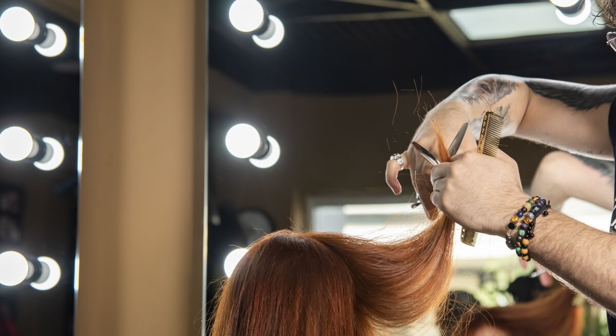 Frau in einem Friseursalon, die gerade ihre langen, roten Haare geschnitten bekommt. Der Frisör hält eine Strähne ihrer Haare und schneidet diese mit einer Schere. Im Hintergrund sind mehrere beleuchtete Spiegel zu sehen.
