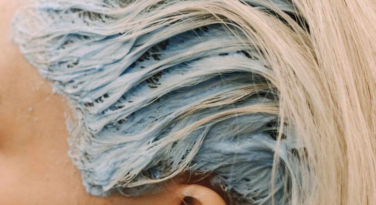 Detail einer Haarfärbung im Prozess. Die blonden Haare sind mit einer blauen Färbemasse bedeckt. Man kann die Struktur der Haare und die Cremigkeit der Farbe erkennen, die einen Kontrast zum natürlichen Blond der Haare bildet.