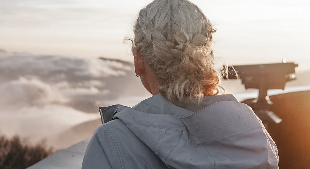 Junge Frau mit grauen, eingeflochtenen Haaren von hinten, die in die Ferne auf eine beeindruckende Berglandschaft blickt.