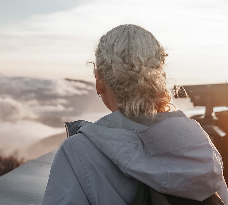 Junge Frau mit grauen, eingeflochtenen Haaren von hinten, die in die Ferne auf eine beeindruckende Berglandschaft blickt. | © Getty Images /	Kendall Haynes / EyeEm