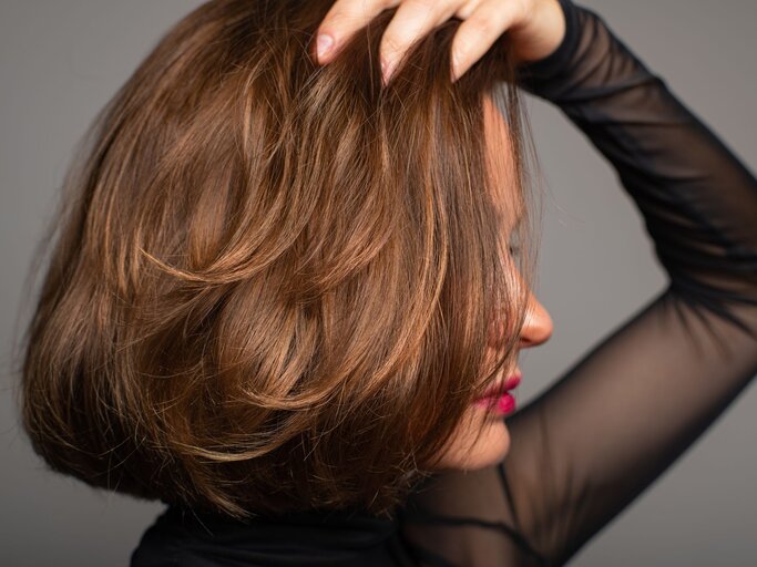 Frau, die mit ihrer Hand durch ihr kurz geschnittenes, voluminöses Haar fährt. Sie trägt einen dunklen, durchsichtigen Ärmel und hat leuchtend roten Lippenstift aufgetragen. Ihr Blick ist vom Betrachter abgewendet | © Adobe Stock / Dmytro Flisak