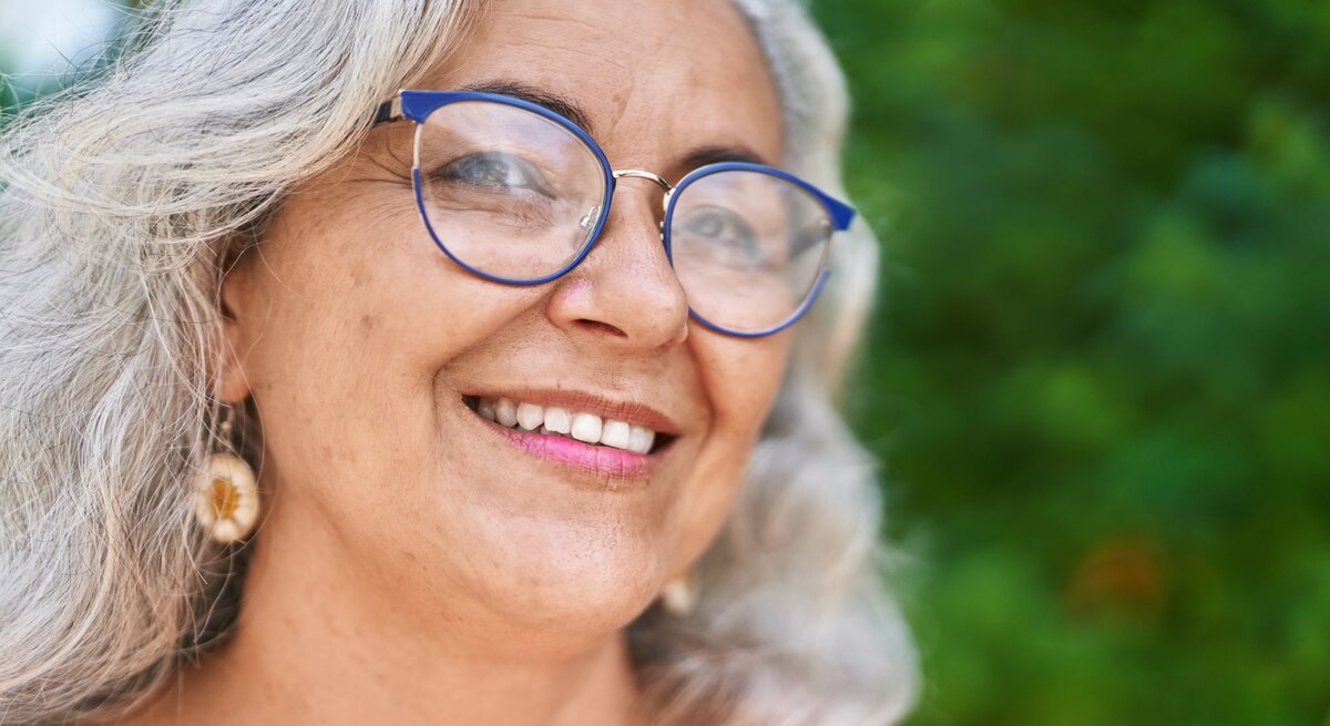 Frau mit grauem Haar, baumelnden Muschel-Ohrringen, einer blauen Brille und rotem Oberteil, umgeben von grünem, unscharfem Naturhintergrund.