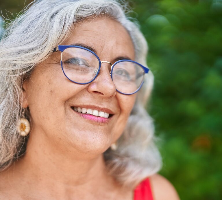 Frau mit grauem Haar, baumelnden Muschel-Ohrringen, einer blauen Brille und rotem Oberteil, umgeben von grünem, unscharfem Naturhintergrund. | © Adobe Stock/ Krakenimages.com