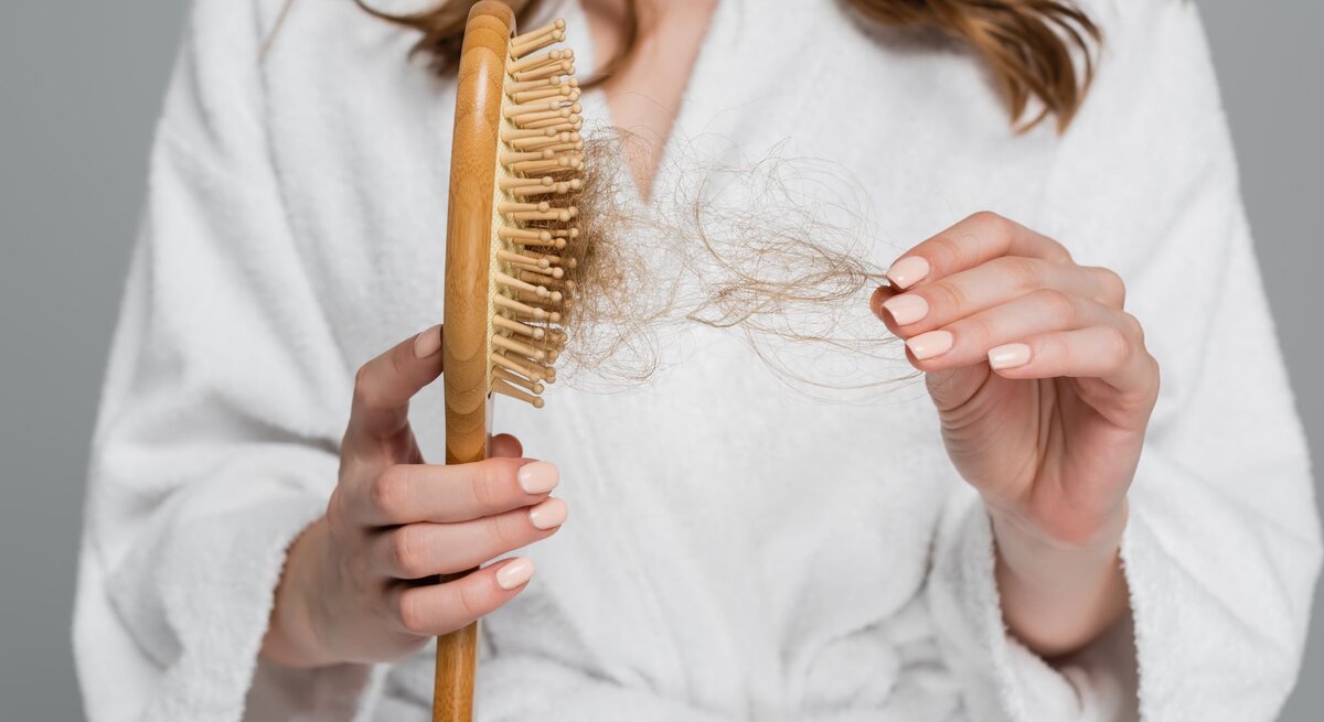 Frau im Bademantel, die besorgt eine Haarbürste mit ausgefallenen Haaren betrachtet. 