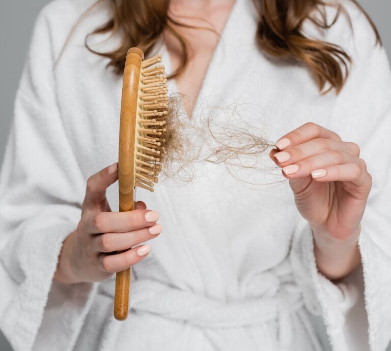 Frau im Bademantel, die besorgt eine Haarbürste mit ausgefallenen Haaren betrachtet.  | © Adobe Stock / LIGHTFIELD STUDIOS