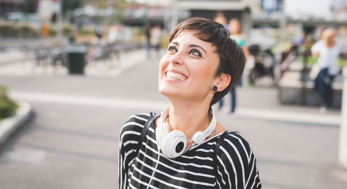 Lächelnde Frau mit einem kurzen Pixie Cut, die draußen steht. Sie trägt ein gestreiftes Oberteil und hat Kopfhörer um den Hals. Ihr Blick ist nach oben gerichtet