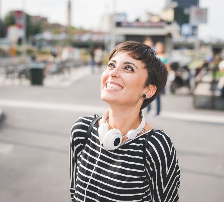 Lächelnde Frau mit einem kurzen Pixie Cut, die draußen steht. Sie trägt ein gestreiftes Oberteil und hat Kopfhörer um den Hals. Ihr Blick ist nach oben gerichtet | © Adobe Stock / Eugenio Marongiu