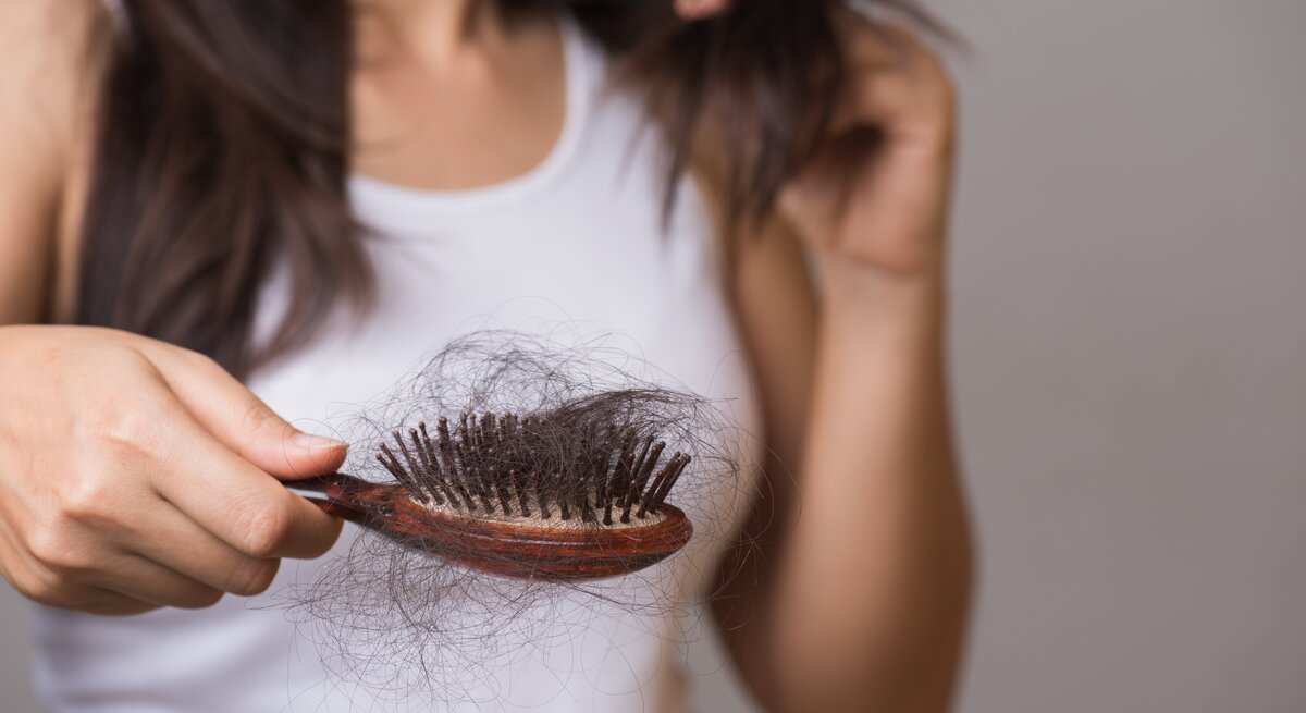 Dunkelhaarige Frau im weißen Top hält besorgt eine Haarbürste voller Haare, während sie eine Haarsträhne prüfend betrachtet.
