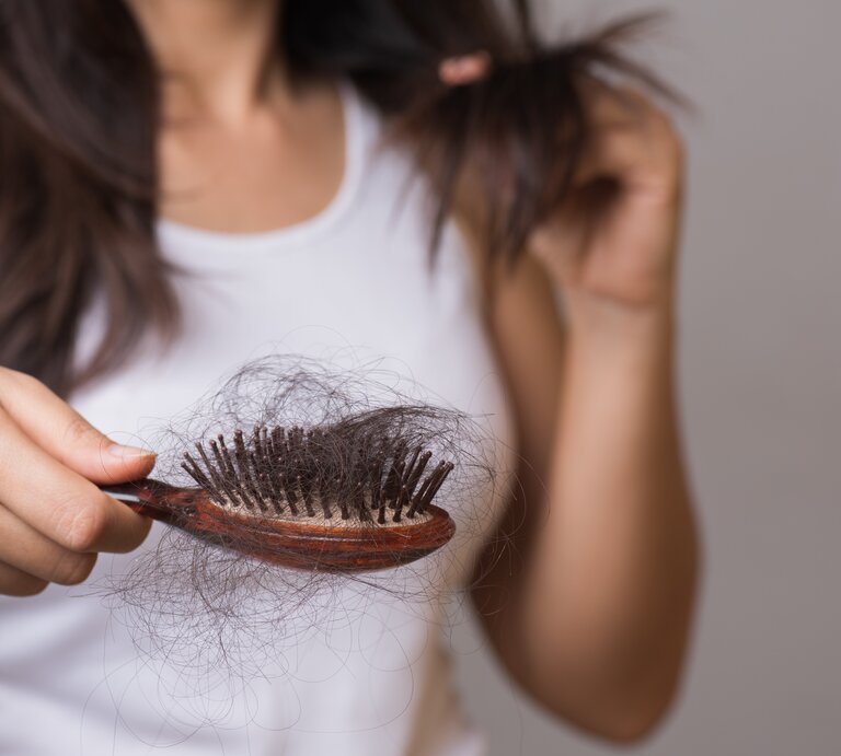 Dunkelhaarige Frau im weißen Top hält besorgt eine Haarbürste voller Haare, während sie eine Haarsträhne prüfend betrachtet. | © Adobe Stock/ Siam