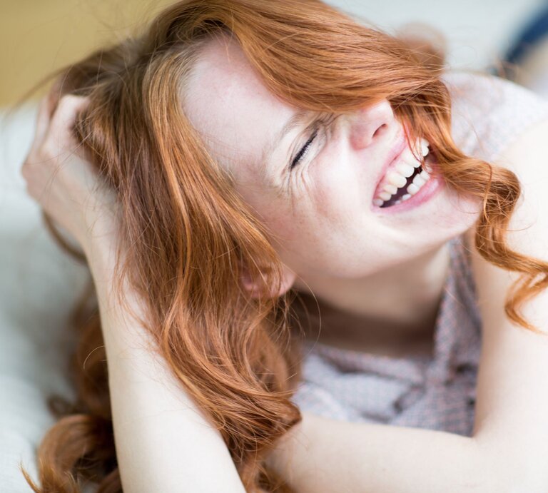 Junge Frau mit langen roten Locken lacht herzlich, während sie sich entspannt auf einem hellen Untergrund hinlegt. | © AdobeStock/drubig-photo