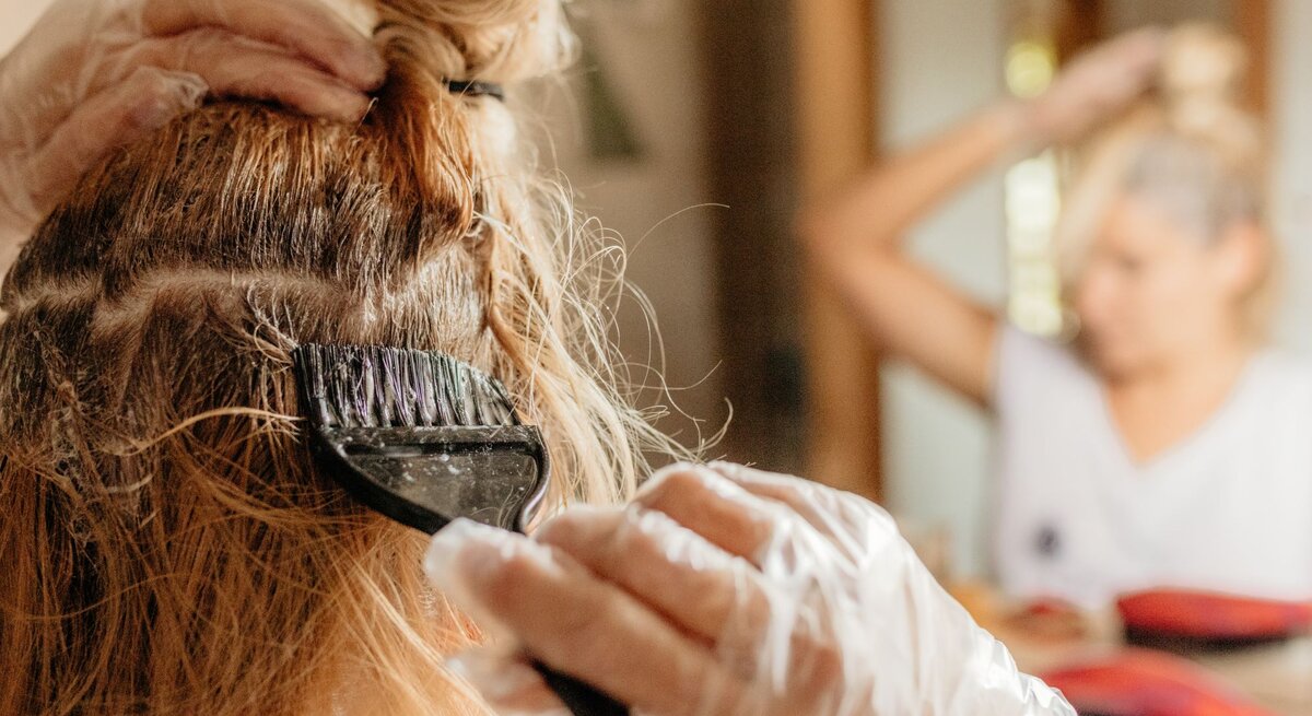 Frau färbt sich ihre Haare vor dem Spiegel mithilfe eines Pinsels, sie trägt Einmalhandschuhe