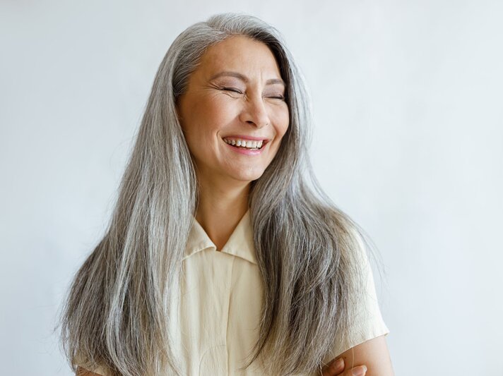 Grauhaarige Frau mittleren Alters in cremefarbener Bluse lacht mit geschlossenen Augen vor hellem, minimalistischem Hintergrund. | © Adobe Stock/ Yaroslav Astakhov