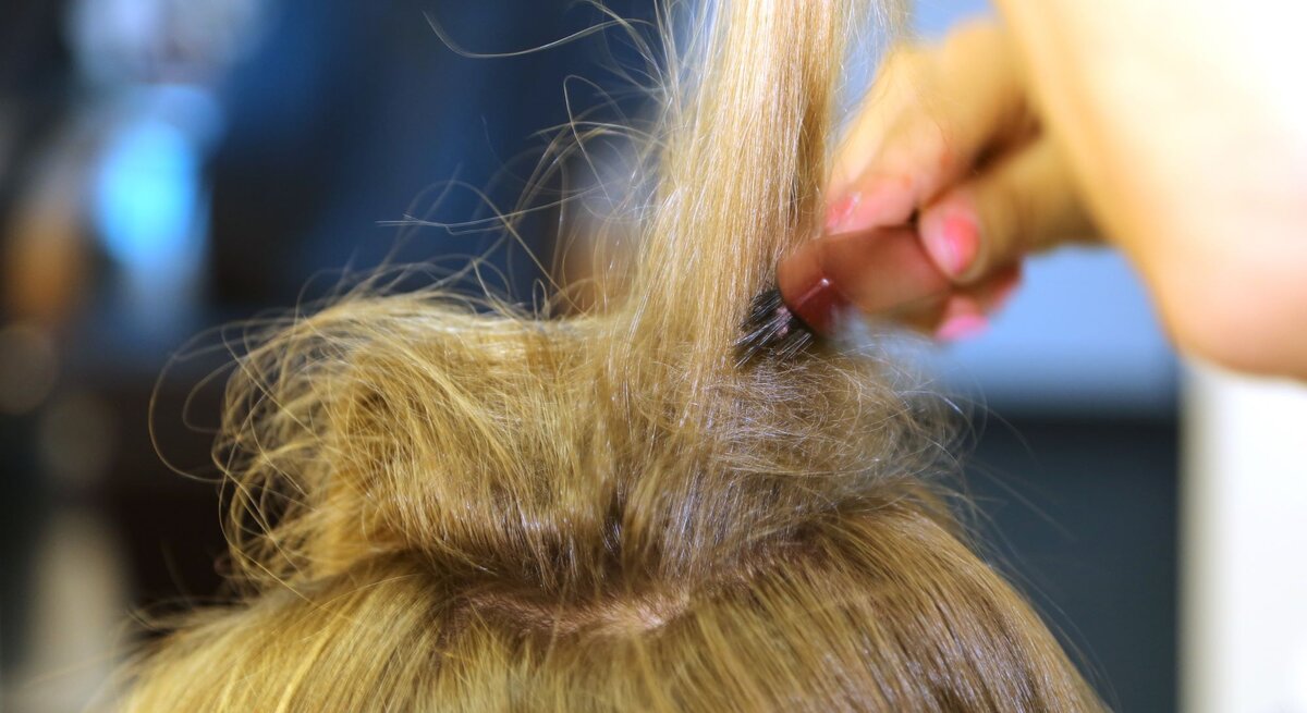 Eine Person toupiert den Haarwirbel auf dem Kopf einer Frau mit langem, blondem Haar, um Volumen zu erzeugen
