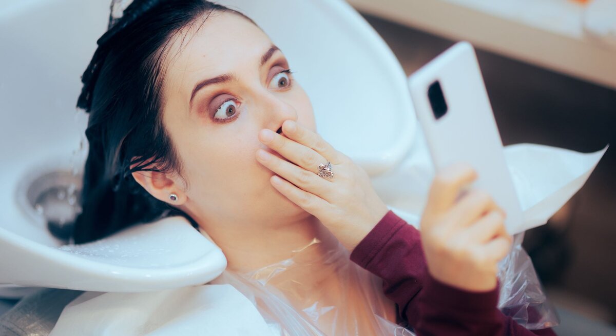 Junge Frau mit dunklem Haar hält schockiert das Handy hoch, während ihr beim Friseur die Haare gewaschen werden.