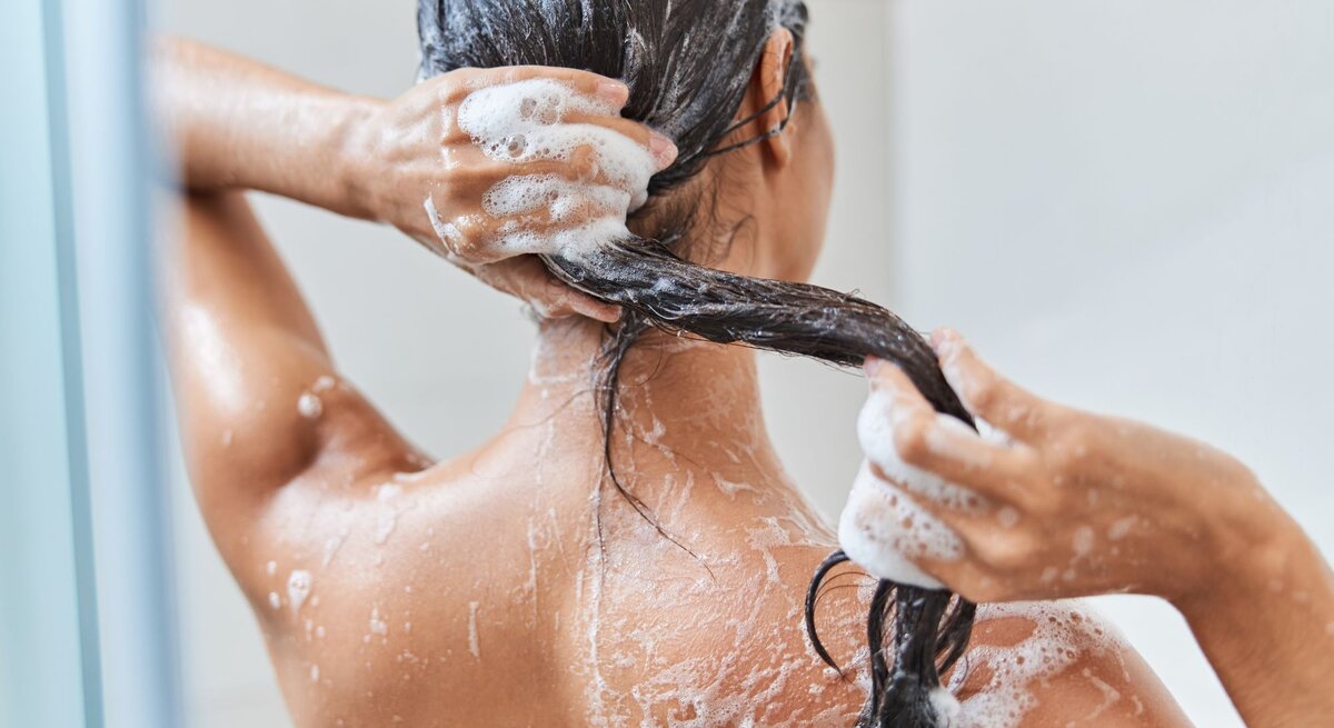 Frau mit nasser Haut schäumt sich unter der Dusche sorgfältig die langen dunklen Haare ein, Rückenansicht im hellen Bad.