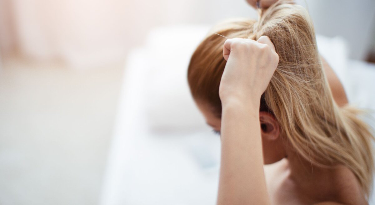 Junge Frau von hinten, die sich gerade die Haare zu einem Pferdeschwanz zusammenbindet. Ihre Haare sind blond.