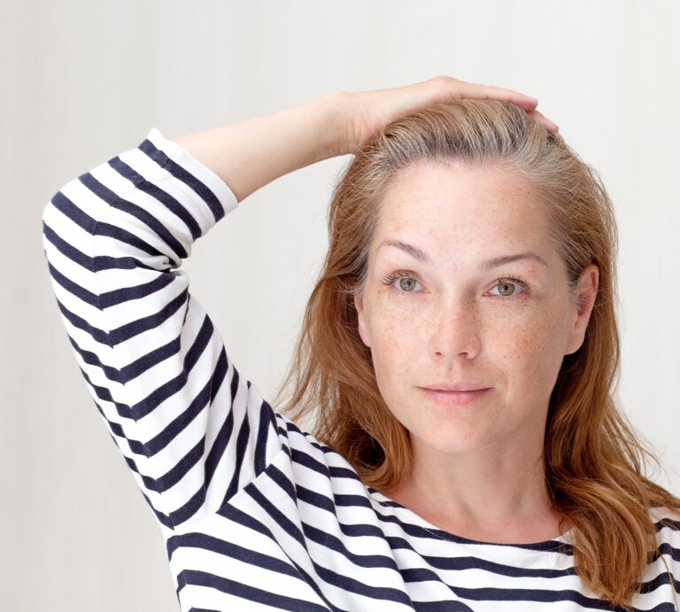 Eine Person mit halblangem rotbraunem Haar und grauem Ansatz trägt ein gestreiftes Shirt und hält eine Hand auf den Kopf vor hellem Hintergrund. | © Adobe Stock/ Tatiana Foxy