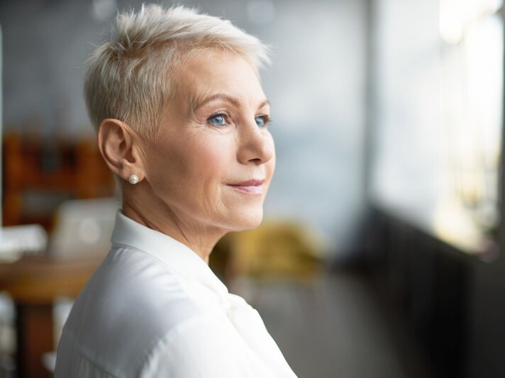 Reife Frau mit stylischer Pixie-Frisur - einer Kurzhaarfrisur für dünnes Haar - und Perlenohrringen trägt weiße Bluse und schaut verträumt ins Tageslicht. | © Adobe Stock/ shurkin_son