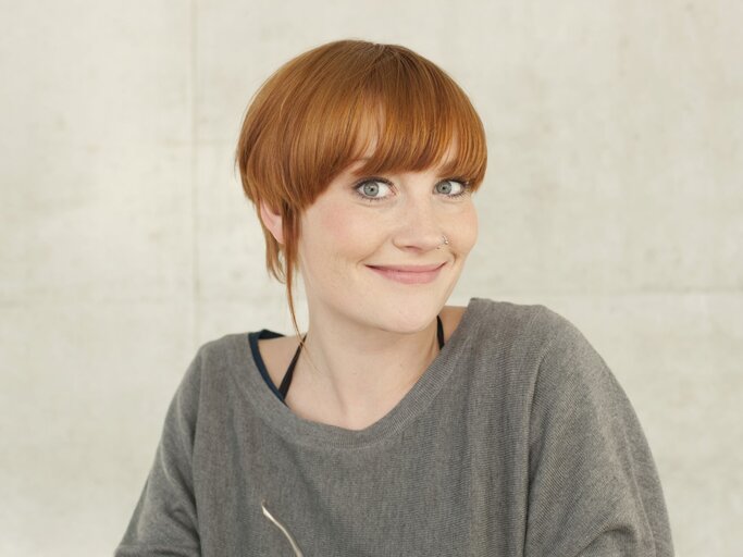 Frau mit leuchtend orangefarbenem, Kurzhaarfrisur und einem sympathischen Lächeln. Sie trägt ein graues Oberteil und hat ein Nasen-Piercing. | © Getty Images / Westend61
