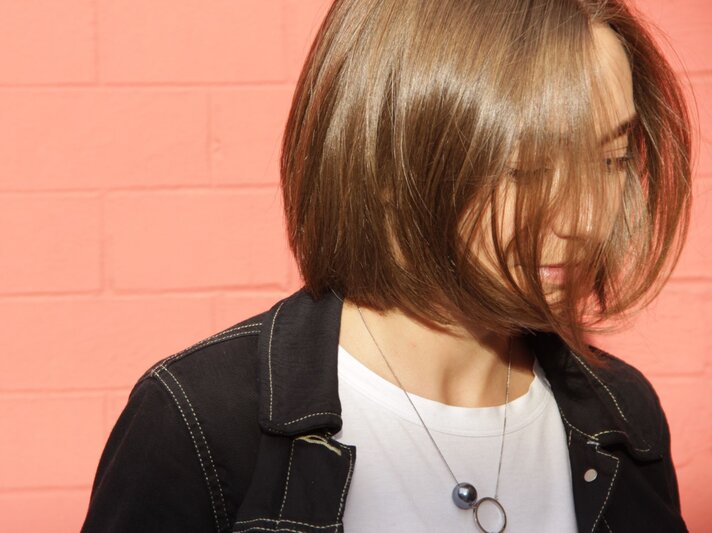 Eine junge Frau mit schulterlangem, braunem Bob trägt ein weißes T-Shirt und eine schwarze Jacke, ihr Blick ist nach unten gerichtet vor pinkem Hintergrund. | © FreePik/ EyeEm