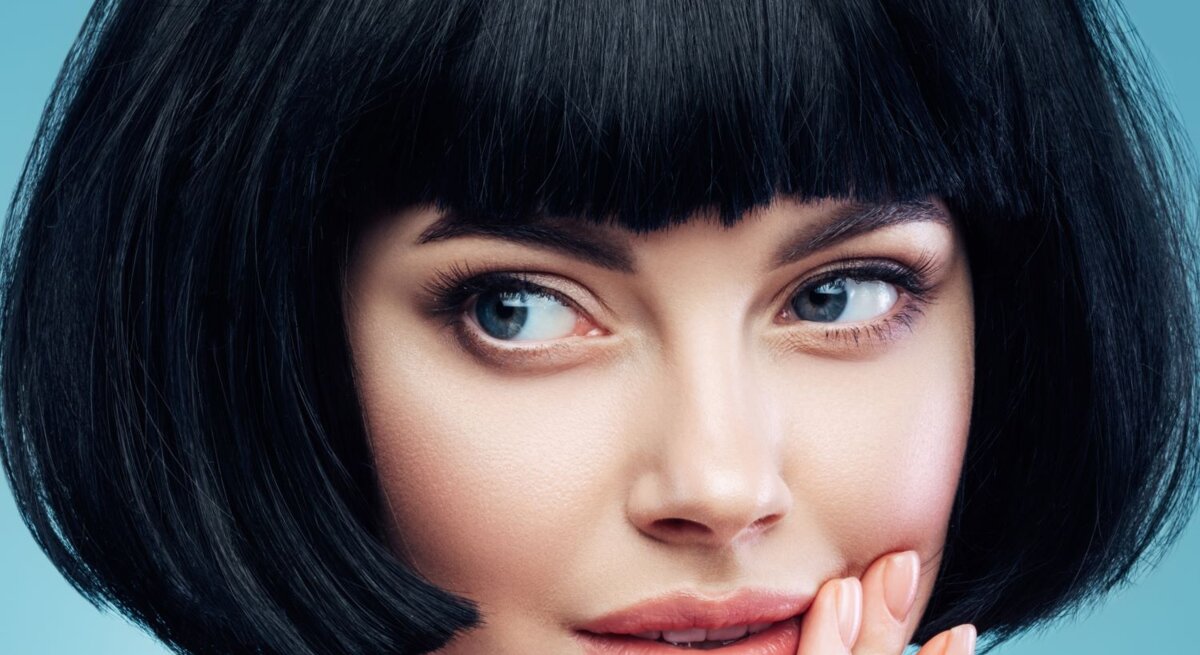 Frau mit einem schwarzen Bob-Haarschnitt und einem blauen Hintergrund. Sie hat blaue Augen und schaut leicht zur Seite, während sie ihre rechte Hand sanft an ihre Wange legt.