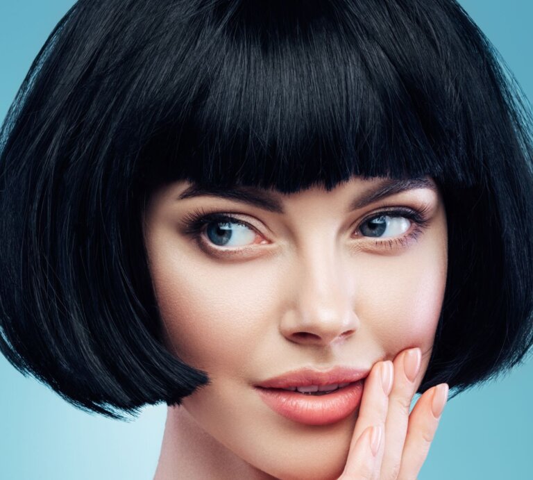 Frau mit einem schwarzen Bob-Haarschnitt und einem blauen Hintergrund. Sie hat blaue Augen und schaut leicht zur Seite, während sie ihre rechte Hand sanft an ihre Wange legt. | © Getty Images / CoffeeAndMilk