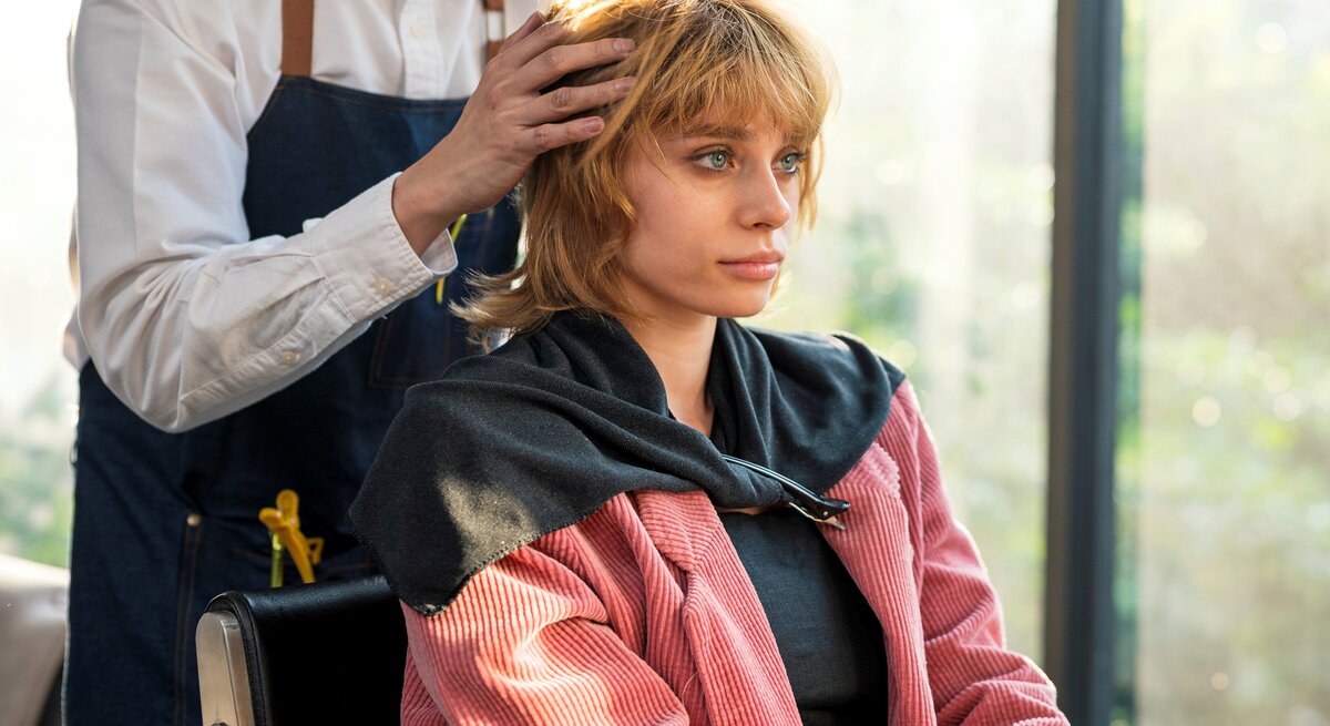 Friseur im weißen Hemd und blauer Schürze stylt Haare einer Kundin in pinkfarbenem Cordmantel vor heller Fensterfront.