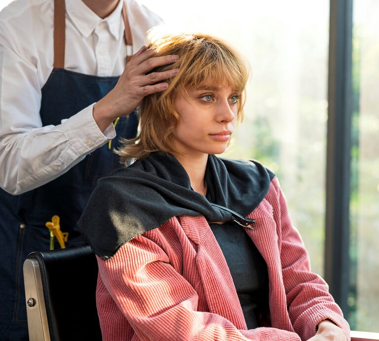 Friseur im weißen Hemd und blauer Schürze stylt Haare einer Kundin in pinkfarbenem Cordmantel vor heller Fensterfront. | © Getty Images / Nitat Termmee