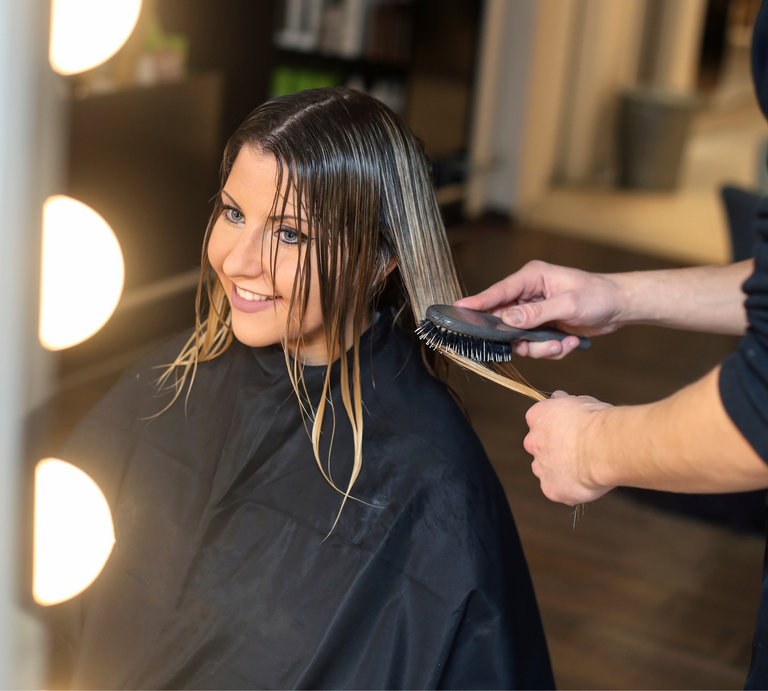 Friseur bürstet blondiertes, nasses Haar einer Person in schwarzem Umhang bei warmem Licht im modernen Salon. | © Canva
