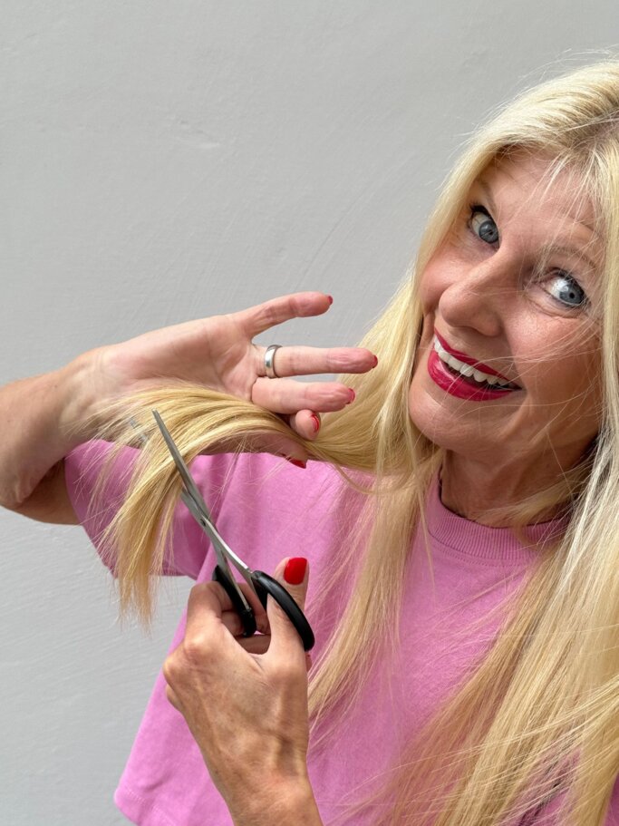 Blondhaarige ältere Frau mit pinkem Lippenstift schneidet lachend eine Haarsträhne mit einer Schere vor grauer Wand. | © Renate Zott