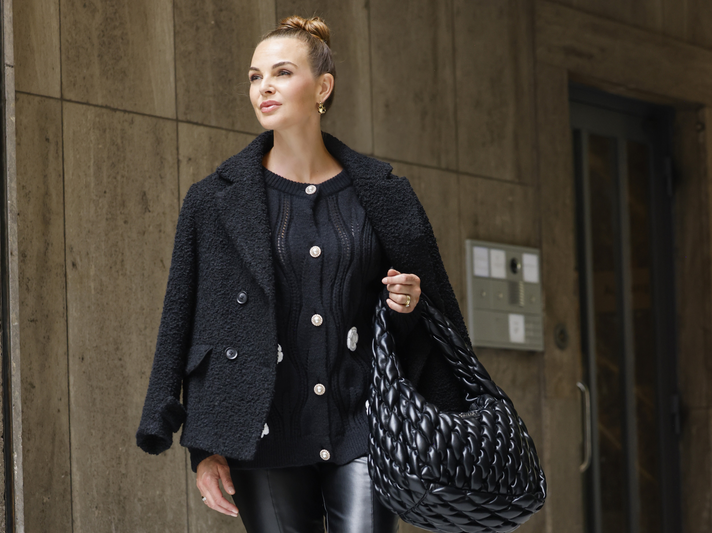Annika Lau in schwarzem Wollmantel und Lederhose trägt eine große geflochtene schwarze Tasche und spaziert vor einer Betonwand. | © Streetstyleshooters for QVC