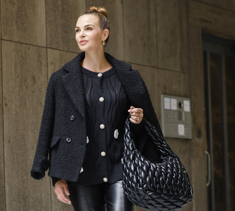 Annika Lau in schwarzem Wollmantel und Lederhose trägt eine große geflochtene schwarze Tasche und spaziert vor einer Betonwand. | © Streetstyleshooters for QVC