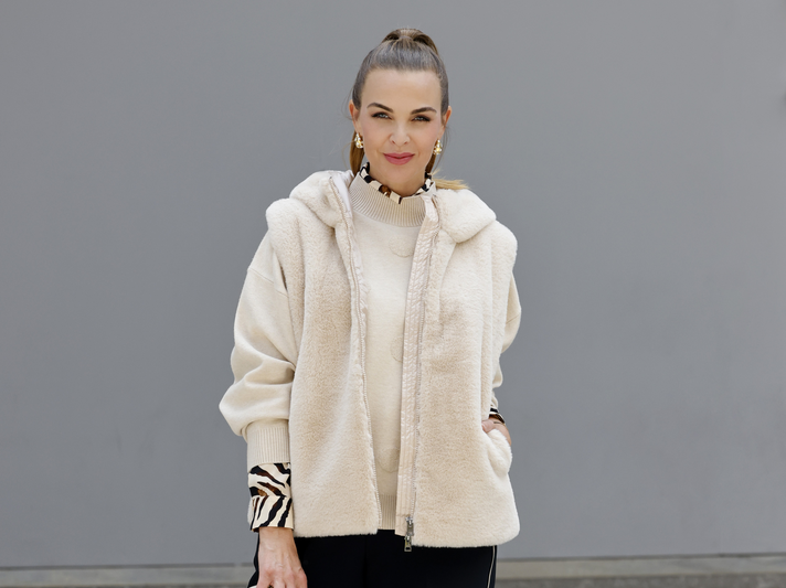 Annika Lau in cremefarbenem Plüschmantel und hellem Rollkragenpullover mit Zebra-Muster-Details steht vor einer grauen Wand. | © Streetstyleshooters for QVC